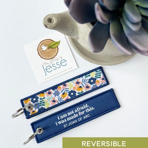 Puede incluir: Dos llaveros rectangulares de tela con anillas met&aacute;licas. Un lado presenta un colorido estampado floral con flores azules, amarillas y rosas sobre fondo blanco. El otro lado es azul marino liso con el texto blanco I am not afraid, I was made for this. ST. JOAN OF ARC.