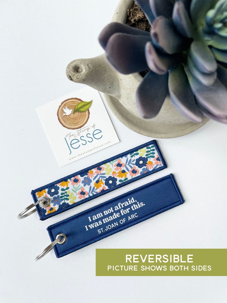 Puede incluir: Dos llaveros rectangulares de tela con anillas met&aacute;licas. Un lado presenta un colorido estampado floral con flores azules, amarillas y rosas sobre fondo blanco. El otro lado es azul marino liso con el texto blanco I am not afraid, I was made for this. ST. JOAN OF ARC.