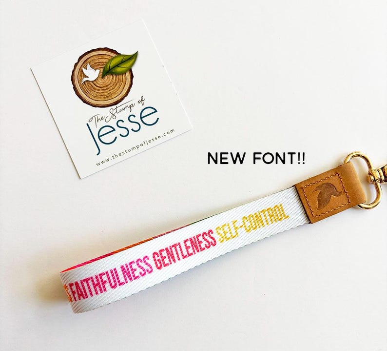 May include: A white fabric wristlet keychain featuring the words FAITHFULNESS, GENTLENESS, and SELF-CONTROL printed in pink and yellow. The strap is attached to a tan leather connector with a gold-toned metal lobster clasp and ring hardware.