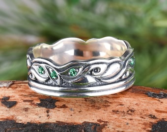 May include: A silver ring with a detailed design of leaves and green gemstones. The ring is set on a wooden surface.