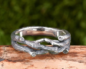 Può includere: Un anello d'argento con un designo di rami intrecciati. L'anello è appoggiato su un pezzo di legno con muschio verde sullo sfondo.