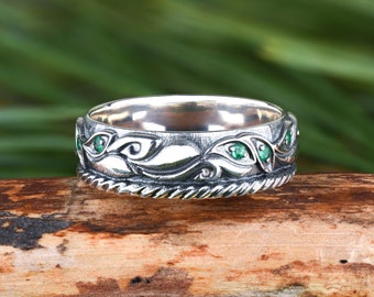 May include: A silver band ring featuring an oxidized finish with embossed swirling leaf patterns and small, round, green gemstones set within the metal. The ring has a twisted rope-style border along the bottom edge of the band.