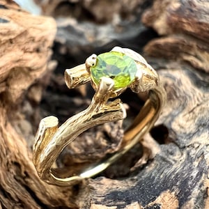 May include: A gold ring featuring a round, faceted, lime-green peridot gemstone set within a band designed to resemble a textured tree branch with small twigs extending upward to hold the stone in a prong setting.