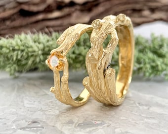 May include: Two gold rings with a textured, organic design resembling tree branches. One ring features a round, light orange gemstone. The rings are displayed on a marble surface with a blurred background of wood and greenery.