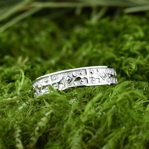 May include: A silver band with an intricate, geometric design. The band is set against a backdrop of green moss.