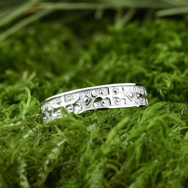 May include: A silver band with an intricate, geometric design. The band is set against a backdrop of green moss.