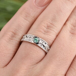 May include: A silver band ring with an intricate design and a small green gemstone in the centre.