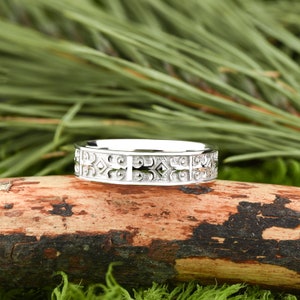 May include: A silver band with a detailed, intricate design. The band is on a wooden surface.