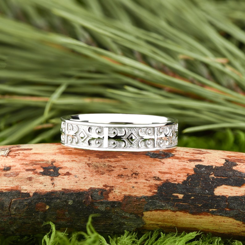 May include: A silver band with a detailed, intricate design. The band is on a wooden surface.