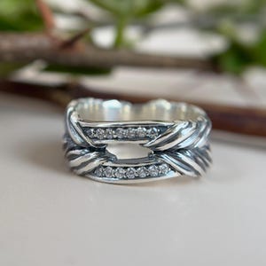 May include: A silver ring with a distinctive design. The ring has a braided pattern and is embellished with small, clear gemstones. This ring is a statement piece, ideal for adding a touch of elegance to any outfit.