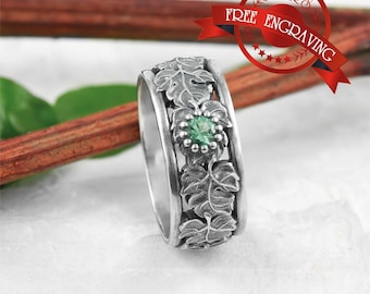 May include: A silver ring with a detailed leaf design and a central green gemstone. The ring features a band with embossed leaves and a cluster of leaves around the gemstone. The ring is displayed on a white surface with a red ribbon that says "FREE ENGRAVING".