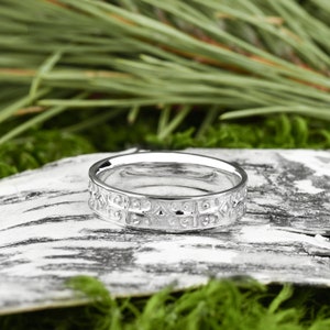 May include: A silver wedding band with a detailed, intricate design. The band is on a white, textured surface.
