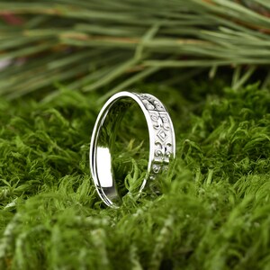 May include: A silver wedding band with a detailed, intricate design. The band is on a bed of green moss.