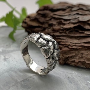 Può includere: Un anello d'argento con un design intrecciato e texture. L'anello è spesso e ha una fascia larga.