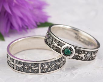 May include: Two silver rings with intricate, dark-toned patterns. One ring features a central green gemstone and small diamonds. The other ring has a similar design. A purple flower is in the background, adding a touch of color.