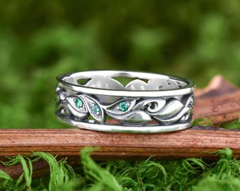 May include: A silver ring with a leaf design and three green gemstones. The ring is set against a backdrop of green moss and a wooden branch, highlighting its natural aesthetic. The ring's intricate details and the vibrant green stones create a striking visual appeal.