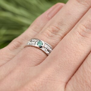 May include: A silver ring featuring a green gemstone set in the centre. The ring has a delicate design with small beads and swirls.