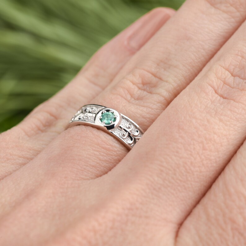 May include: A silver ring featuring a green gemstone set in the centre. The ring has a delicate design with small beads and swirls.