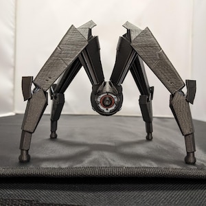 May include: A dark grey, mechanical spider robot with six articulated legs and a central body. The robot has a detailed design with visible textures and a red and black eye-like component. The legs are angled, giving the robot a dynamic pose.