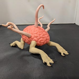 Op de afbeelding: Een grillige sculptuur van een roze hersenvorm met lichtbruine ledematen en tentakels. De sculptuur heeft een getextureerd oppervlak en staat tegen een donkere achtergrond.