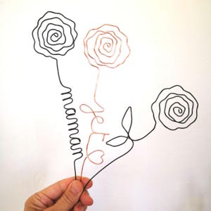 Könnte beinhalten: Draht-Skulptur aus drei Rosen, eine Rose ist kupferfarben und die anderen beiden sind schwarz. Die kupferne Rose hat das Wort "Love" auf dem Stiel geschrieben. Die schwarze Rose links hat das Wort "Maman" auf dem Stiel geschrieben.