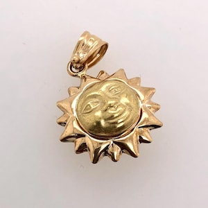 May include: A gold pendant charm in the shape of a sun with a smiling face. The sun has a textured surface and is surrounded by rays.