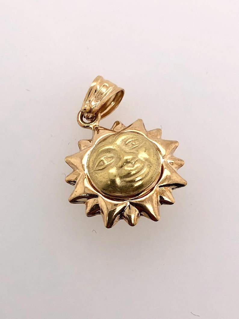 May include: A gold pendant charm in the shape of a sun with a smiling face. The sun has a textured surface and is surrounded by rays.