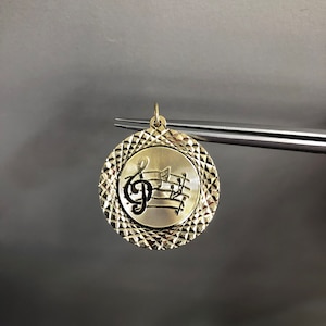 May include: A gold-colored pendant with a treble clef and musical notes engraved on a round, textured surface.