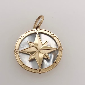 May include: Gold compass pendant with a white mother of pearl inlay. The compass features a gold star with four points and the letters N, S, E, and W around the edge.