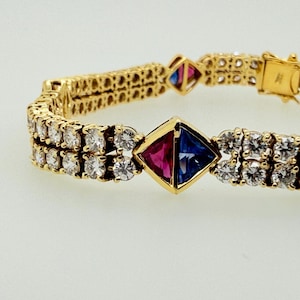 May include: A gold bracelet with two rows of small, clear gemstones. The bracelet features square-shaped accents with red and blue gemstones. The clasp is gold and the bracelet is on a white background.