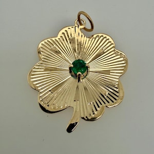 May include: A gold-colored four-leaf clover pendant with a small, round, green gemstone in the center. The pendant has a textured, ridged design radiating from the center. A small loop is at the top for a chain.