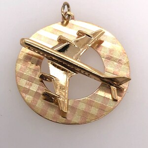 May include: A gold-toned pendant featuring a stylized airplane design. The airplane is set against a circular background with a textured pattern.