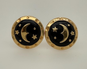 May include: A pair of gold-toned clip-on earrings. Each earring features a black enamel center with a gold crescent moon and stars design. The earrings have a decorative gold border.