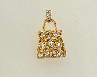 May include: A gold-colored pendant in the shape of a handbag, adorned with small, sparkling diamonds. The handbag design features an openwork pattern, and the pendant has a small loop at the top for a chain.