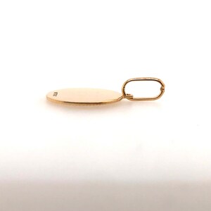 Pode incluir: Um pingente de ouro com um berloque circular e uma argola retangular. O pingente &eacute; de uma cor dourada amarela quente e parece ser feito de metal. O berloque &eacute; liso e plano, com uma pequena marca na superf&iacute;cie. A argola &eacute; presa ao topo do berloque.