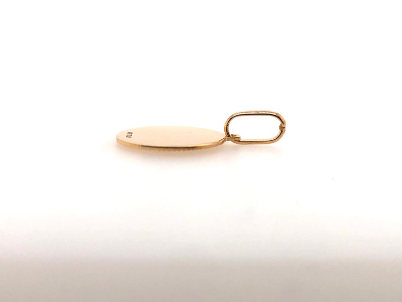Pode incluir: Um pingente de ouro com um berloque circular e uma argola retangular. O pingente &eacute; de uma cor dourada amarela quente e parece ser feito de metal. O berloque &eacute; liso e plano, com uma pequena marca na superf&iacute;cie. A argola &eacute; presa ao topo do berloque.