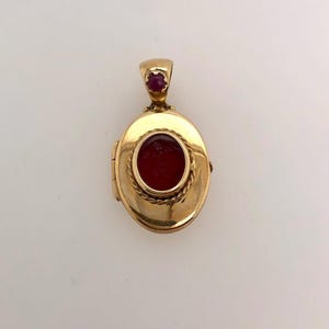 May include: A gold oval locket with a red stone in the center and a smaller red stone on the top. The locket has a rope-like design around the edge.