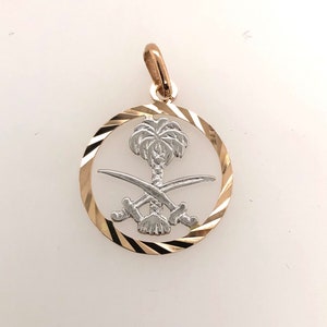 May include: A gold pendant with a circular design featuring a white palm tree and two crossed swords. The pendant has a textured edge.