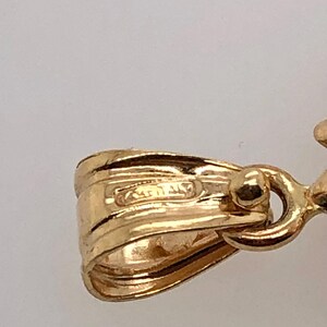 May include: Close-up of a gold pendant with a detailed design. The pendant has a loop for attaching to a chain and a small, round bead on the side.