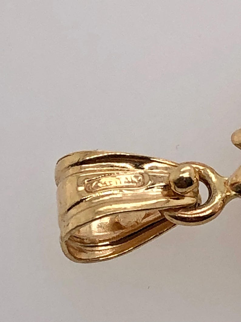 May include: Close-up of a gold pendant with a detailed design. The pendant has a loop for attaching to a chain and a small, round bead on the side.