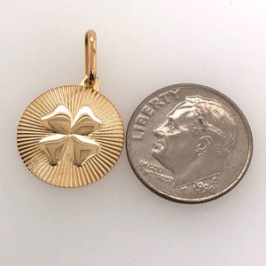Pode incluir: Pingente dourado com um design de trevo de quatro folhas. O pingente redondo apresenta um padr&atilde;o texturizado em forma de raios de sol ao redor do trevo. Uma moeda est&aacute; ao lado para compara&ccedil;&atilde;o de tamanho. O di&acirc;metro &eacute; de aproximadamente 2 cm.