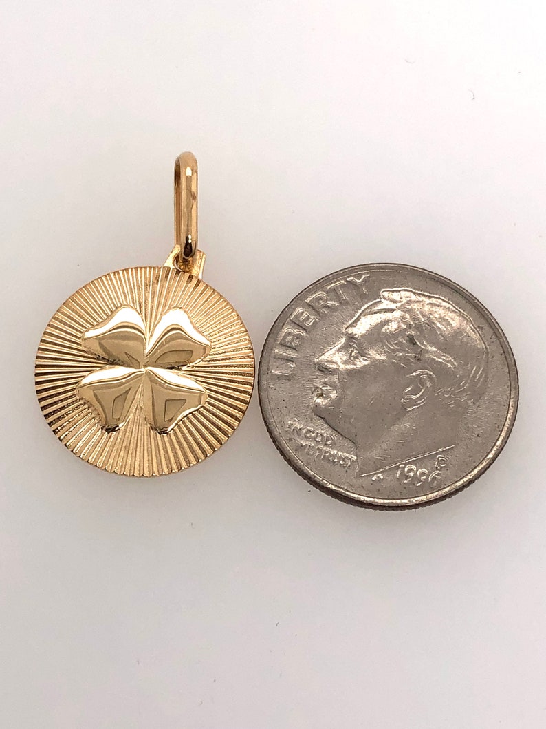 Pode incluir: Pingente dourado com um design de trevo de quatro folhas. O pingente redondo apresenta um padr&atilde;o texturizado em forma de raios de sol ao redor do trevo. Uma moeda est&aacute; ao lado para compara&ccedil;&atilde;o de tamanho. O di&acirc;metro &eacute; de aproximadamente 2 cm.