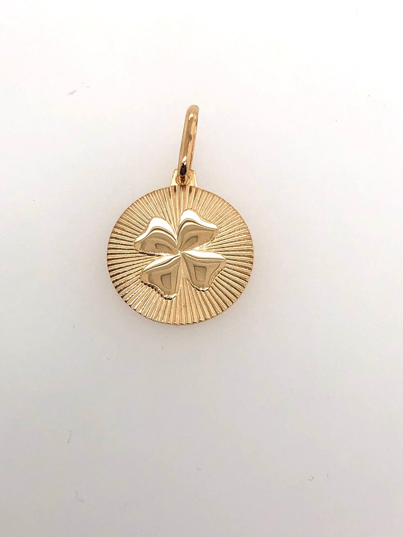Pode incluir: Pingente dourado com um design de trevo de quatro folhas. O trevo est&aacute; centrado em um disco circular com um padr&atilde;o de raios de sol. O pingente inclui um pequeno la&ccedil;o para prender a uma corrente ou pulseira.