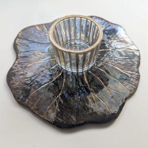 Könnte beinhalten: Ein Kerzenhalter aus Keramik mit einem durchsichtigen Glasbecher, der auf einer blattförmigen Basis steht. Die Basis ist dunkel mit goldenen Akzenten, und der Glasbecher hat vertikale Linien. Der Kerzenhalter ist dekorativ.