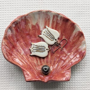 May include: A ceramic seashell-shaped dish in shades of red and pink holds a pair of white tulip earrings with silver hooks. The earrings have gold and gray accents. A small, dark gray pearl sits in the center of the dish.