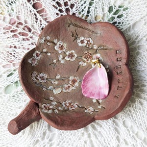 May include: A handmade, brown ceramic dish with a floral design and a pink pendant. The dish features a branch with white flowers. The pendant is a light pink petal shape with a gold-colored bail. The dish is on a white lace surface.