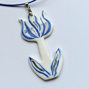 May include: A white ceramic pendant shaped like a tulip flower with blue painted details and metallic silver accents. The pendant hangs from a thin blue cord attached by a silver-toned metal bail, creating a decorative piece of jewelry.