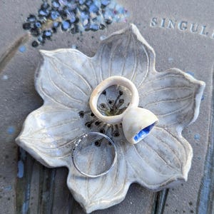 Può includere: Un portagioie in ceramica a forma di fiore nei toni del grigio e del bianco, con tre anelli. Un anello è d'argento, gli altri due sono in ceramica, uno con l'interno blu. Il piatto è su una superficie strutturata con accenti floreali.