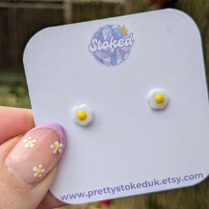 May include: A pair of white and yellow fried egg earrings on a white card with the text "Stoked" and "www.prettystokeduk.etsy.com".