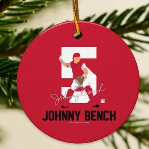 May include: A red, round Christmas ornament featuring a baseball player illustration. The number 5 is prominently displayed, with the name "JOHNNY BENCH" below. A gold string is attached for hanging. The ornament is set against a backdrop of evergreen branches.